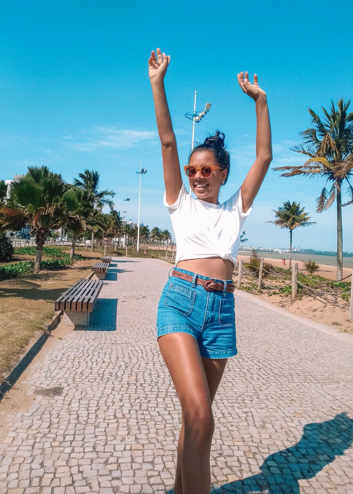 Mulher sorridente de braços levantados, usando camiseta branca e shorts jeans, caminha no calçadão de Camburi, um dos melhores lugares onde ficar em Vitória.