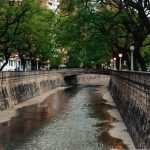 Vista do canal La Cañada em Córdoba com pouca água fluindo e ladeado por muros de pedra e árvores verdes. Postes com luminárias acompanham as calçadas dos dois lados do canal.