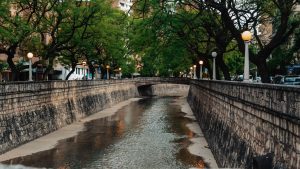 Vista do canal La Cañada em Córdoba com pouca água fluindo e ladeado por muros de pedra e árvores verdes. Postes com luminárias acompanham as calçadas dos dois lados do canal.