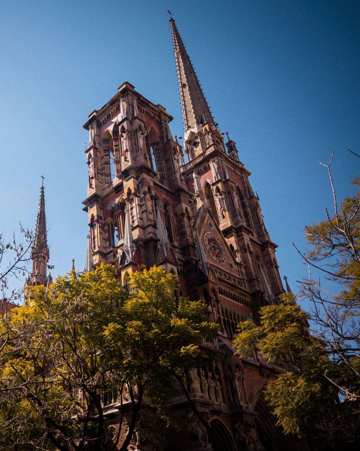 nova-cordoba-argentina-2 Igreja em Nova Córdoba, o melhor lugar para ficar em Córdoba, com sua arquitetura neogótica detalhada, torres altas e vitrais coloridos contra um céu azul intenso. Árvores com folhas amarelas parcialmente escondem a base da construção.