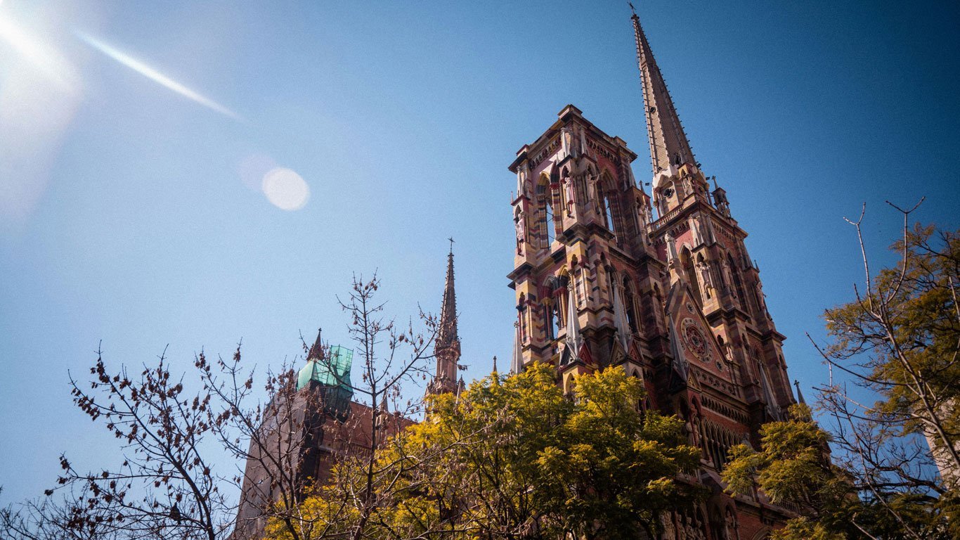 nueva-cordoba-argentina Igreja em Nova Córdoba com sua arquitetura neogótica detalhada, torres altas e vitrais coloridos contra um céu azul intenso. Árvores com folhas amarelas parcialmente escondem a base da construção.