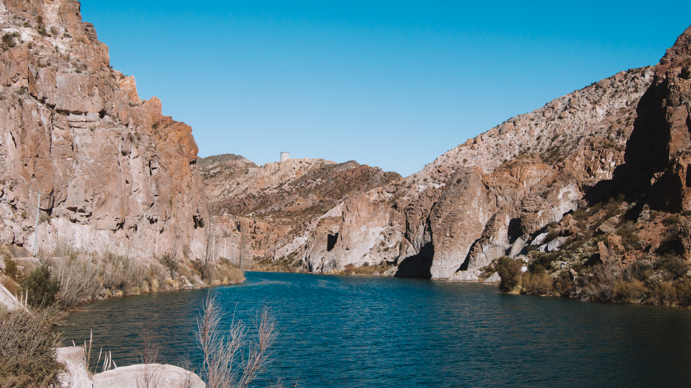 o-que-fazer-em-mendoza-em-novembro Cañón del Atuel com águas azul-escuras serpenteando entre montanhas áridas e íngremes de pedras avermelhadas em Mendoza. Vegetação rasteira cresce nas margens do rio sob o céu claro.