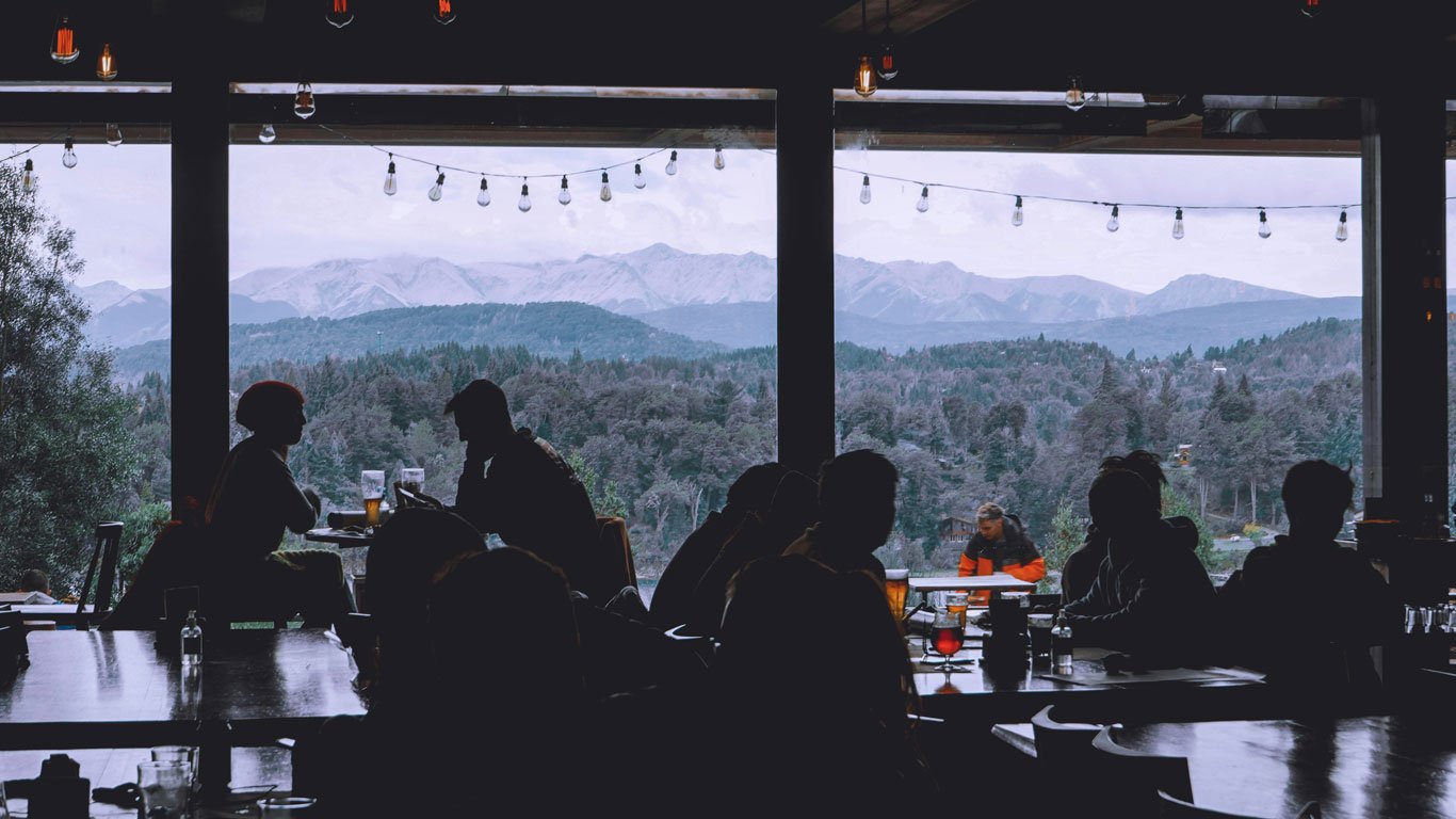 onde-comer-em-bariloche-na-primavera Interior de um restaurante aconchegante com grandes janelas panorâmicas, onde pessoas conversam e apreciam bebidas enquanto observam uma vista deslumbrante das montanhas e florestas de Bariloche em novembro.
