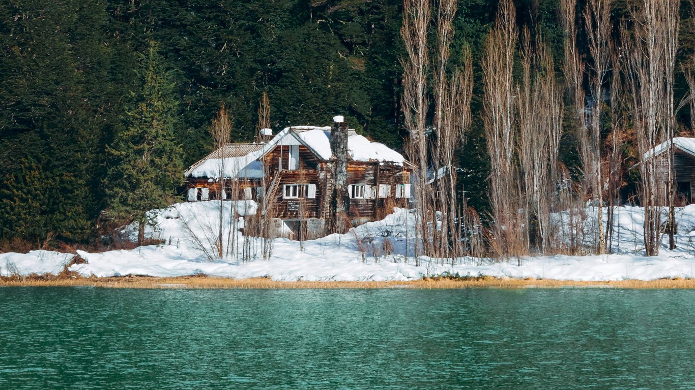 Cabana de madeira coberta de neve cercada por árvores altas e densas em frente a um lago de águas esverdeadas em Bariloche.