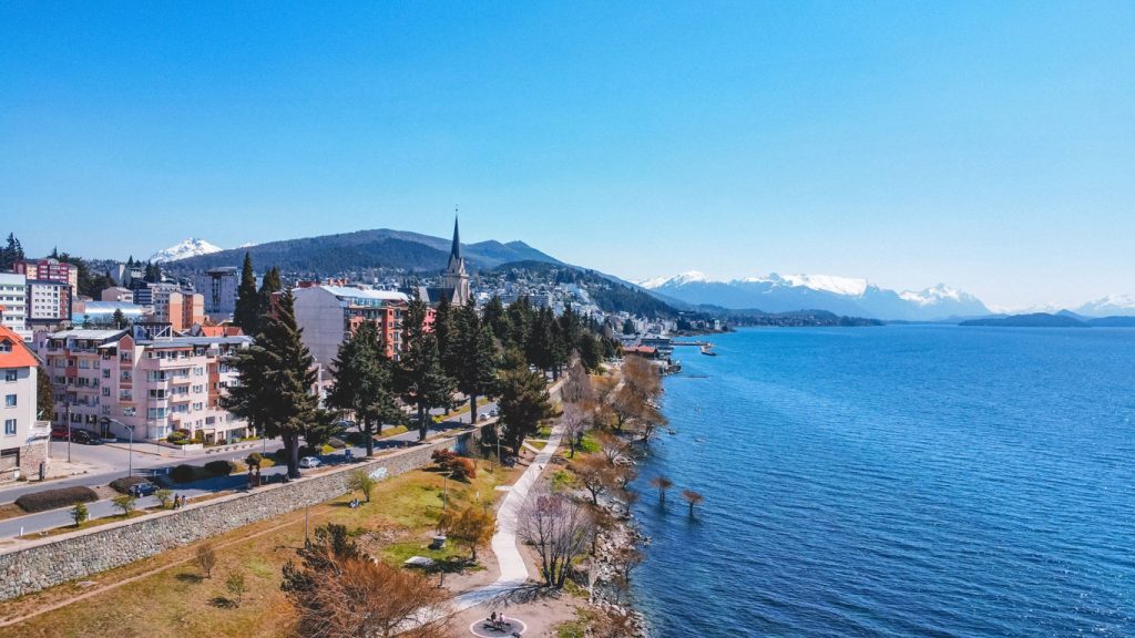 Vista aérea do centro de Bariloche, mostrando edifícios coloridos, uma igreja com torre alta e um calçadão à beira do Lago Nahuel Huapi com montanhas nevadas ao fundo.