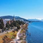Vista aérea do centro de Bariloche, mostrando edifícios coloridos, uma igreja com torre alta e um calçadão à beira do Lago Nahuel Huapi com montanhas nevadas ao fundo.