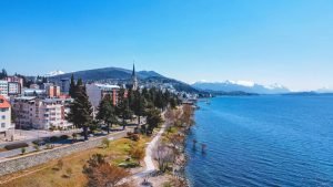 Vista aérea do centro de Bariloche, mostrando edifícios coloridos, uma igreja com torre alta e um calçadão à beira do Lago Nahuel Huapi com montanhas nevadas ao fundo.