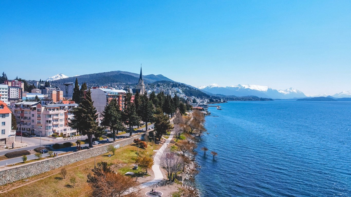 Vista aérea do centro de Bariloche, mostrando edifícios coloridos, uma igreja com torre alta e um calçadão à beira do Lago Nahuel Huapi com montanhas nevadas ao fundo.