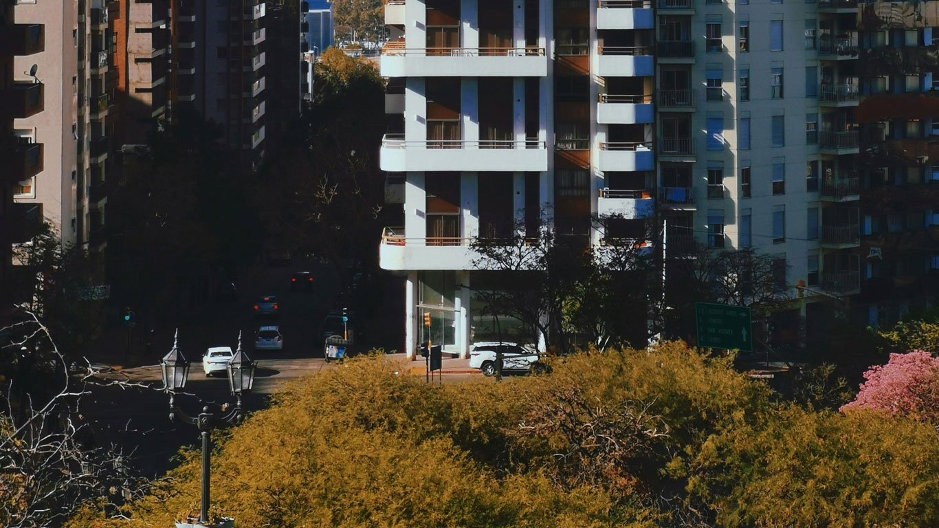 onde-ficar-em-cordoba-argentina1 Imagem de edifícios residenciais altos em uma rua sombreada de Córdoba, vista de cima com árvores douradas em primeiro plano. Carros circulam na rua ao fundo entre os prédios.