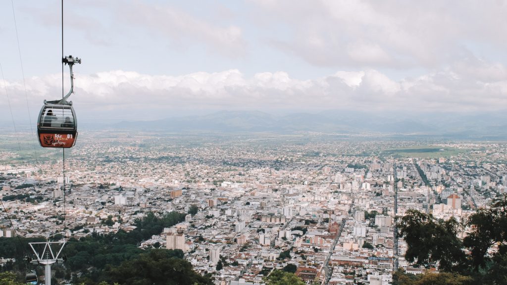Imagem de Salta vista de um mirante com um bondinho em primeiro plano. A cidade se funde com o céu meio nublado no horizonte.