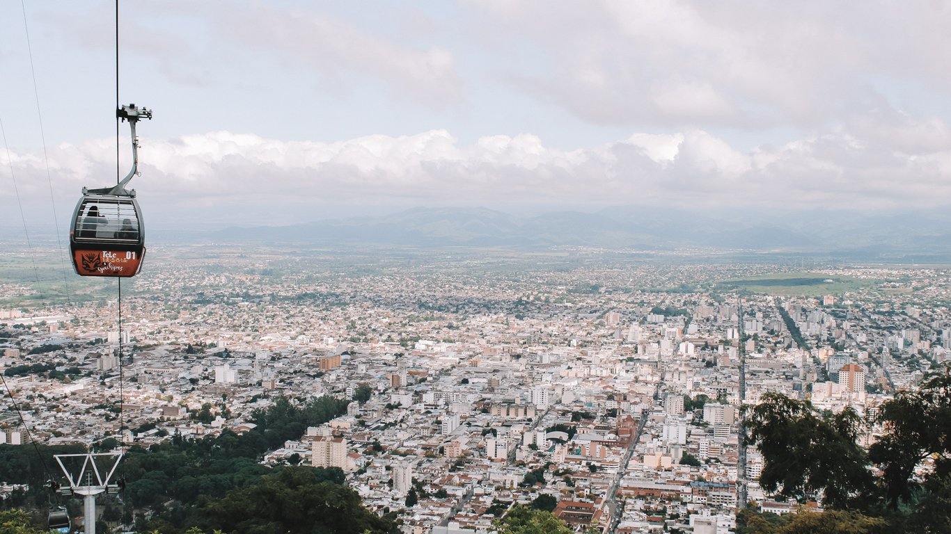 onde-ficar-em-salta-centro Imagem de Salta vista de um mirante com um bondinho em primeiro plano. A cidade se funde com o céu meio nublado no horizonte.