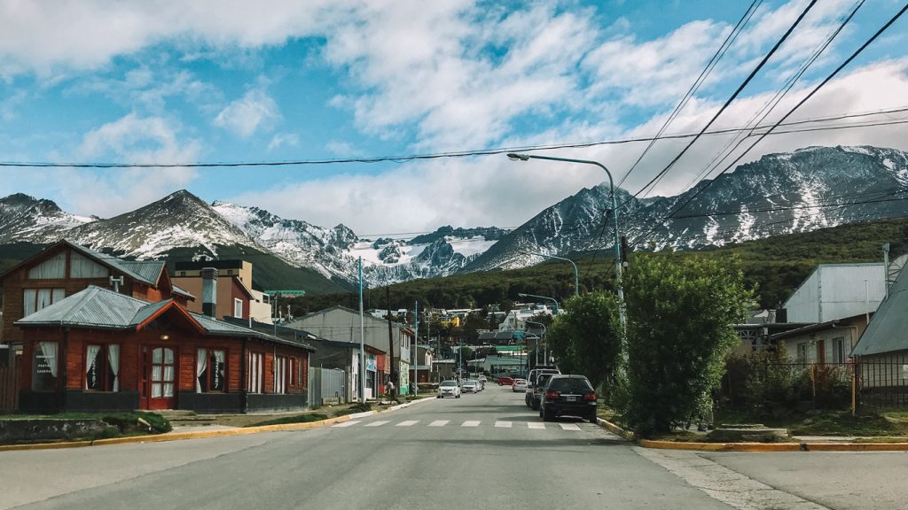 Rua tranquila no centrinho, um dos melhores lugares onde ficar em Ushuaia, com casas de madeira e carros estacionados, cercada por montanhas parcialmente cobertas de neve sob um céu azul com nuvens dispersas.
