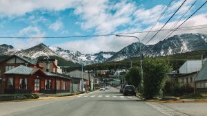 Rua tranquila no centrinho, um dos melhores lugares onde ficar em Ushuaia, com casas de madeira e carros estacionados, cercada por montanhas parcialmente cobertas de neve sob um céu azul com nuvens dispersas.