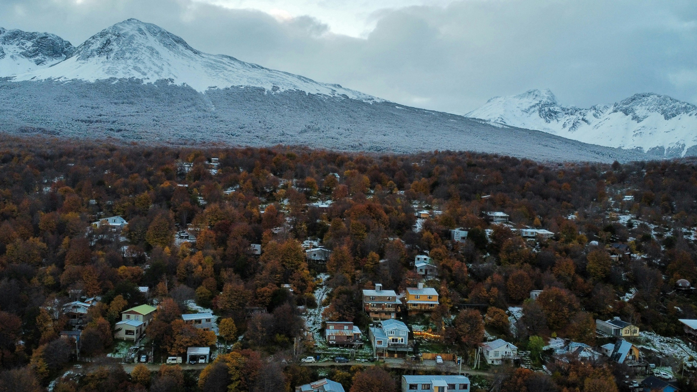 onde-ficar-em-ushuaia-arredores-do-centro Vista aérea de um bairro residencial próximo do centrinho de Ushuaia, em meio a uma floresta de árvores em tons de outono, com montanhas nevadas ao fundo cobrindo a paisagem.