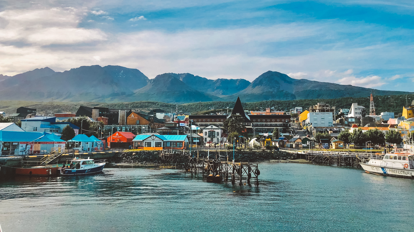 onde-ficar-em-ushuaia-centro Vista do porto de Ushuaia com casas coloridas, barcos atracados e montanhas ao fundo; o céu parcialmente nublado destaca a arquitetura charmosa da cidade.