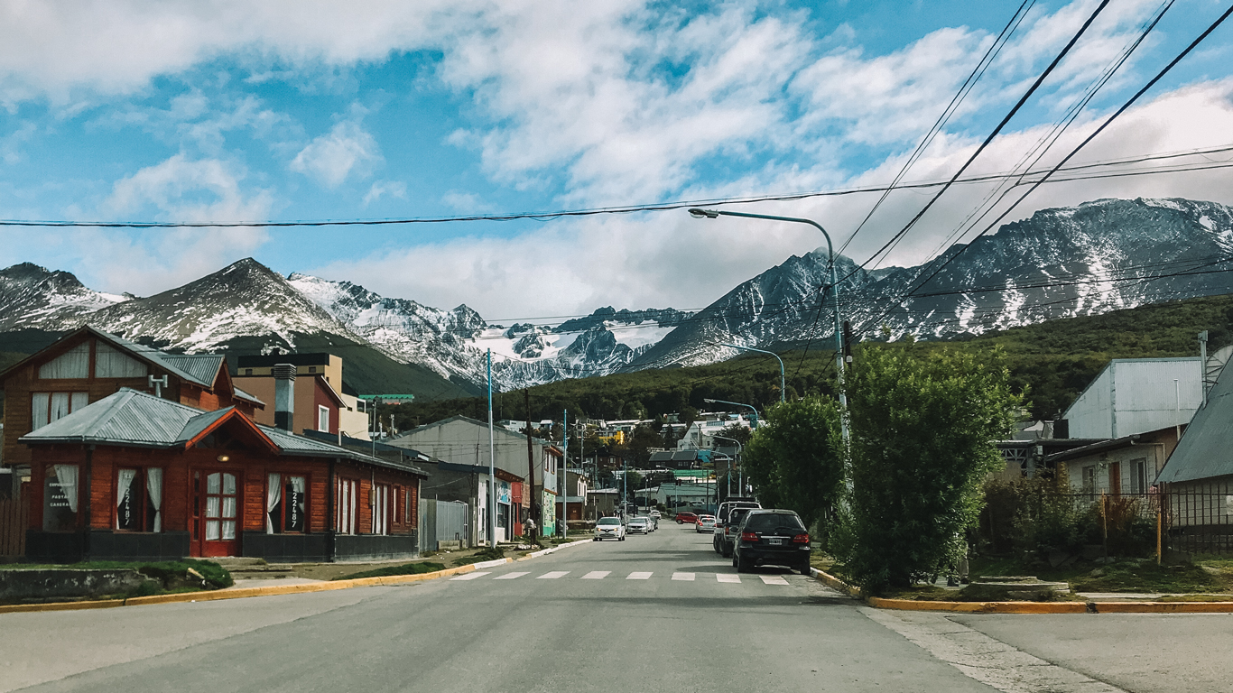 Rua tranquila no centrinho, um dos melhores lugares onde ficar em Ushuaia, com casas de madeira e carros estacionados, cercada por montanhas parcialmente cobertas de neve sob um céu azul com nuvens dispersas.
