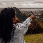 Mulher de costas com cabelo longo preso em um rabo de cavalo, vestindo uma jaqueta jeans clara, aponta para o horizonte em um mirante com vista panorâmica de uma cidade rodeada por morros verdes e mar ao fundo. O céu está nublado e o cenário transmite um clima contemplativo em meio à natureza.