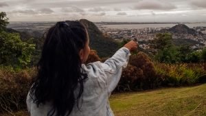 Mulher de costas com cabelo longo preso em um rabo de cavalo, vestindo uma jaqueta jeans clara, aponta para o horizonte em um mirante com vista panorâmica de uma cidade rodeada por morros verdes e mar ao fundo. O céu está nublado e o cenário transmite um clima contemplativo em meio à natureza.