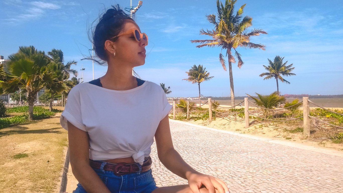 Mulher sentada de perfil em um calçadão à beira da Praia de Camburi, usando óculos de sol, camiseta branca e shorts jeans, com palmeiras e mar ao fundo.
