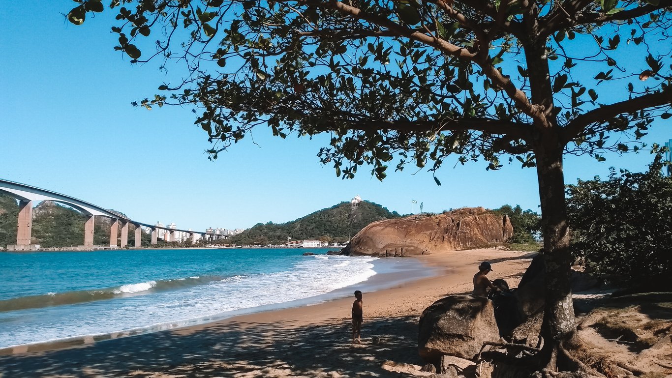Cena tranquila de uma praia na Enseada do Suá em Vitória com criança na areia, homem à sombra de uma árvore, mar calmo, terceira ponte e Convento da Penha ao fundo.