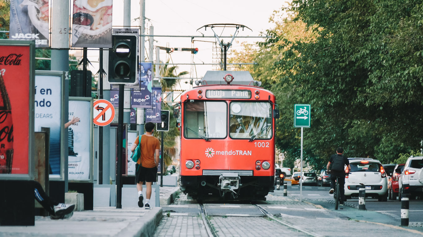 paisagens-mendoza-em-novembro Bonde vermelho da linha "mendoTRAN" circulando pelas ruas de Mendoza com ciclistas, pedestres e carros ao redor. Placas de trânsito, outdoors e árvores margeiam a via.