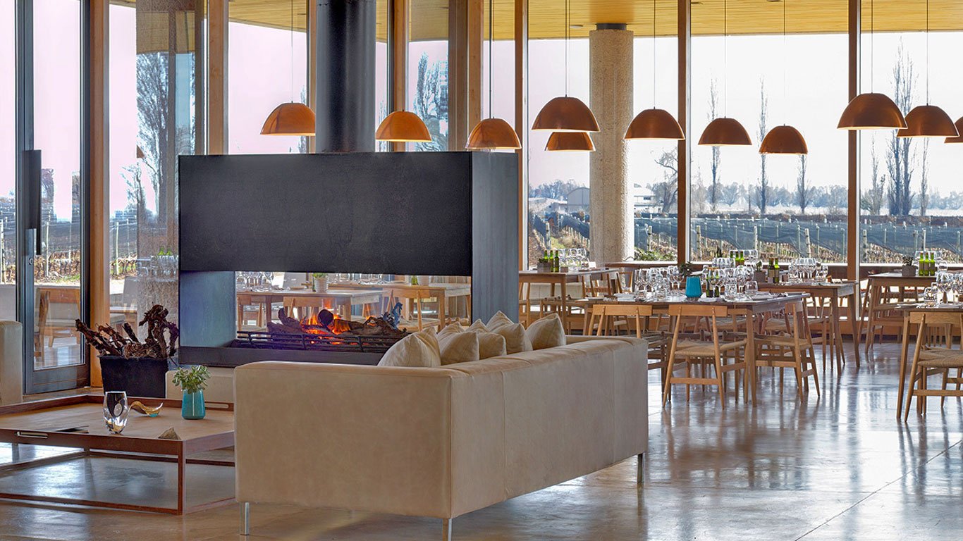 restaurante-zucardi Ambiente interno elegante e aconchegante da vinícola Zuccardi em Mendoza, com lareira moderna no centro, sofás claros e mesas de jantar arrumadas com taças e garrafas. Grandes janelas de vidro oferecem vista panorâmica dos vinhedos ao fundo, iluminados pela luz natural.
