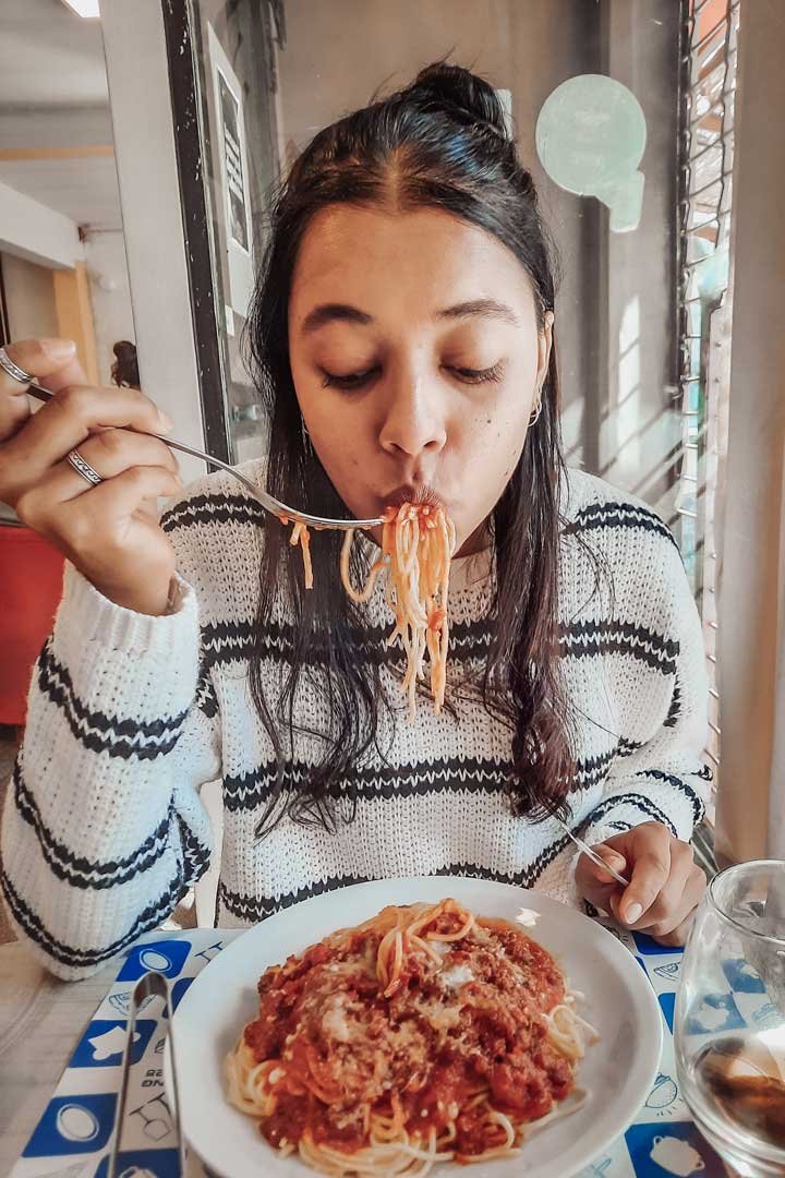 restaurantes-em-mendoza-5 Mulher com suéter listrado comendo um prato de espaguete com molho vermelho e queijo ralado, sugando os fios com um garfo. Ela está sentada à mesa de um restaurante de Mendoza, com um copo de água ao lado, apreciando a refeição de olhos fechados.