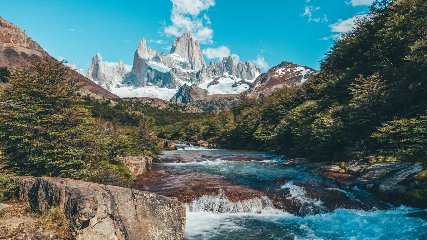 torres-del-paine Rio de águas claras correndo por entre rochas e árvores verdes, com as Torres del Paiine imponente ao fundo em um dia ensolarado com céu azul.