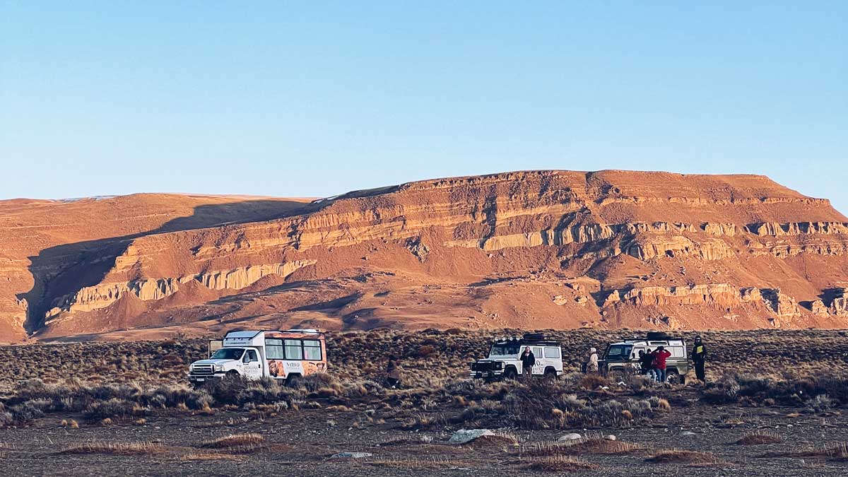 tour-4x4-el-calafate Veículos 4x4 e turistas em uma paisagem de estepa patagônica nos Balcones de Calafate, com formações rochosas avermelhadas iluminadas pela luz dourada do entardecer.