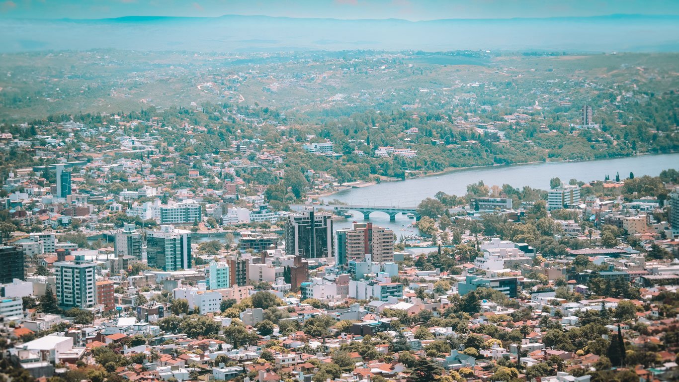 villa-carlos--paz-cordoba Panorama de Villa Carlos Paz com muitos prédios, casas e áreas verdes espalhadas até as montanhas ao fundo. O Rio San Antonio corta a cidade com uma ponte visível no centro da imagem.