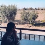 Mulher sentada em uma varanda da vinícola Vistalba, umas das melhores vinícolas em Mendoza com almoço, olhando pensativa para um vinhedo ensolarado em Mendoza, com oliveiras ao longo da cerca e o horizonte marcado por árvores e pequenas construções.