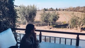 Mulher sentada em uma varanda da vinícola Vistalba, umas das melhores vinícolas em Mendoza com almoço, olhando pensativa para um vinhedo ensolarado em Mendoza, com oliveiras ao longo da cerca e o horizonte marcado por árvores e pequenas construções.