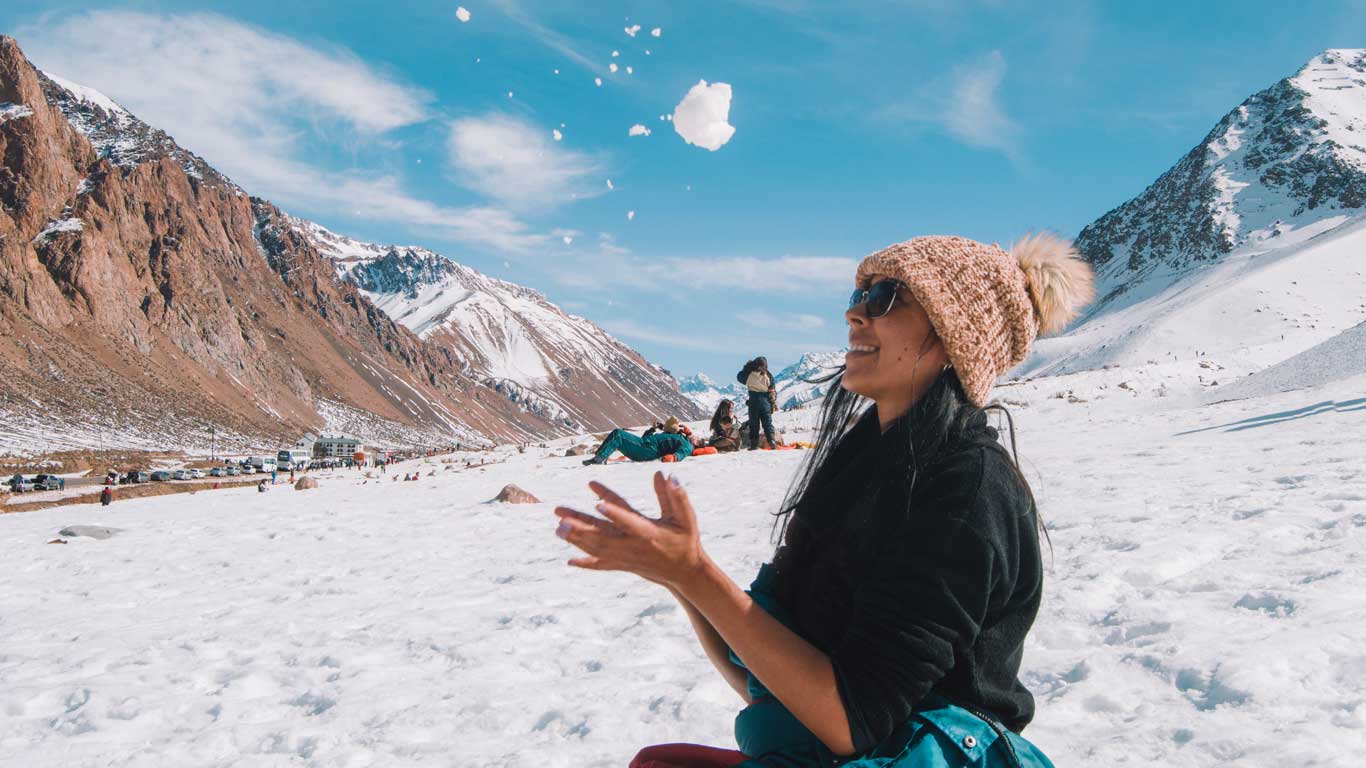 Mulher sorridente, usando gorro de lã com pompom e óculos de sol, brinca jogando neve para o alto em um passeio de inverno na Alta Montanha em Mendoza. Ao fundo, montanhas cobertas parcialmente de neve e céu azul claro.
