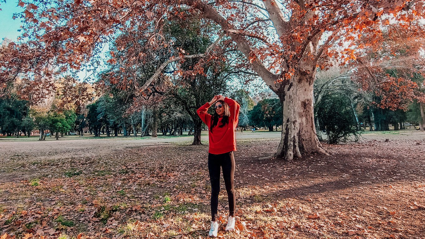 Mulher de moletom vermelho, calça preta e tênis brancos posa debaixo de uma grande árvore com folhas alaranjadas de outono, no Parque General San Martin em Mendoza, Argentina. O chão está coberto por folhas secas e o sol ilumina suavemente a cena.