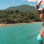 Mulher olhando uma praia isolada em Ilhabela, cercada por vegetação, com casas de veraneio em sua orla. O mar esverdeado com águas cristalinas complementa a paisagem.