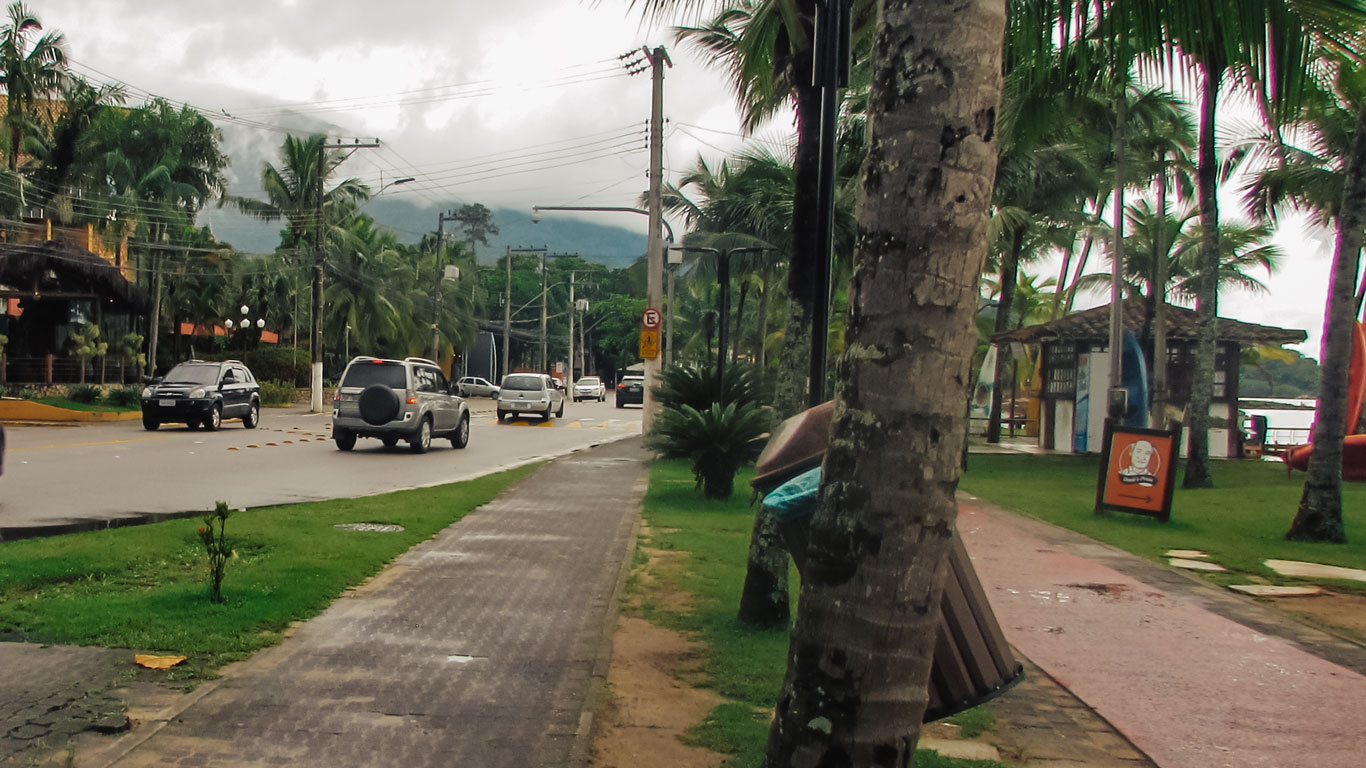 Rua movimentada na Praia do Perequê, uma das principais regiões onde ficar em Ilhabela com crianças, com calçada larga, ciclovia e palmeiras ao longo da orla. Do lado direito, há um quiosque de aluguel de pranchas e caiaques voltado para o mar, enquanto carros circulam pela avenida em frente à área comercial.