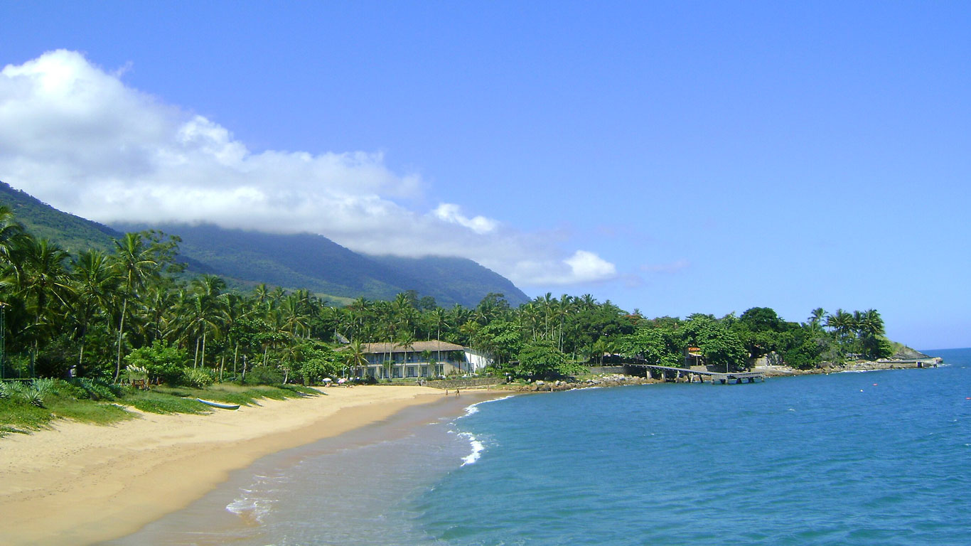 Praia da Feiticeira em Ilhabela com areia dourada, mar calmo e águas azuladas. Ao fundo, colinas verdes cobertas por vegetação tropical e nuvens baixas parcialmente encobrindo o topo das montanhas.