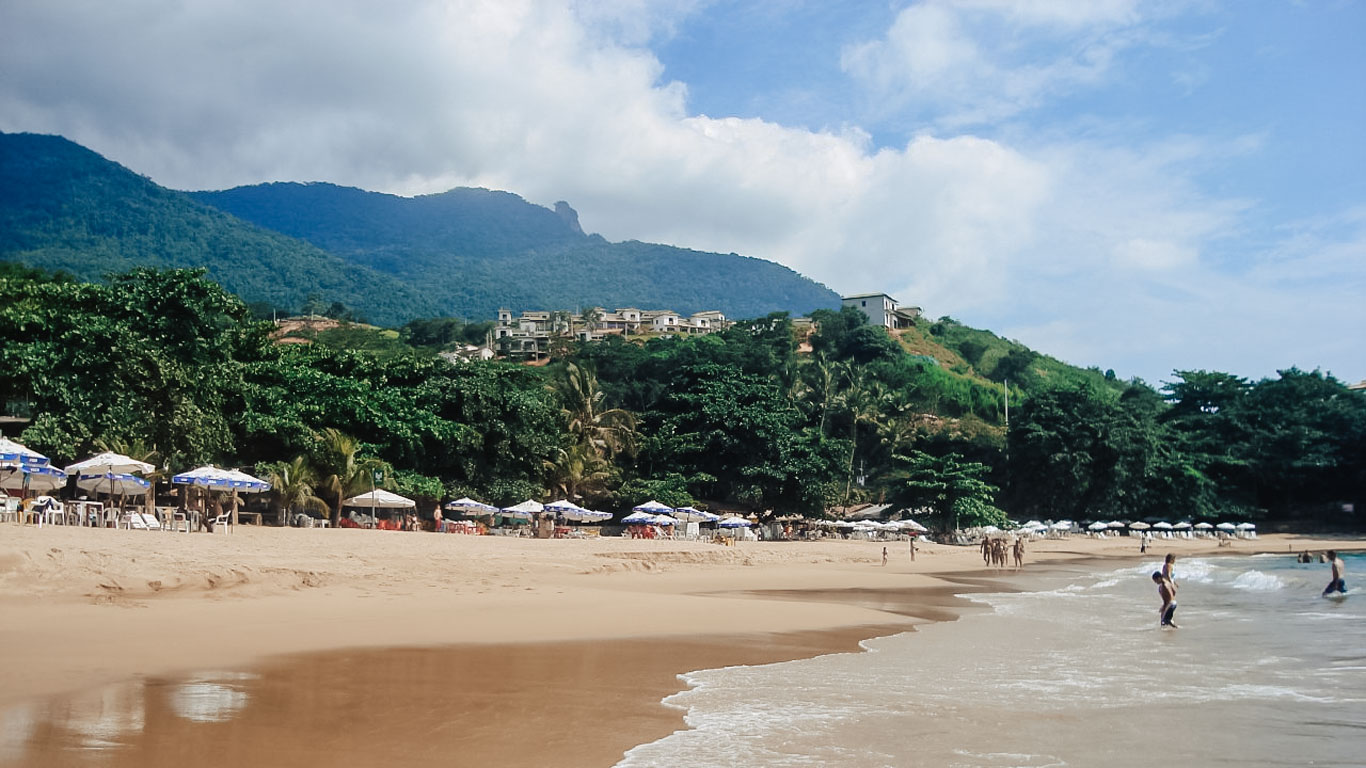 Praia do Curral em Ilhabela, um dos melhores lugares onde ficar em Ilhabela, com areia dourada, mar calmo e guarda-sóis alinhados ao longo da faixa de areia. Ao fundo, morros cobertos por mata atlântica e casas construídas na encosta, com vista privilegiada para o mar.