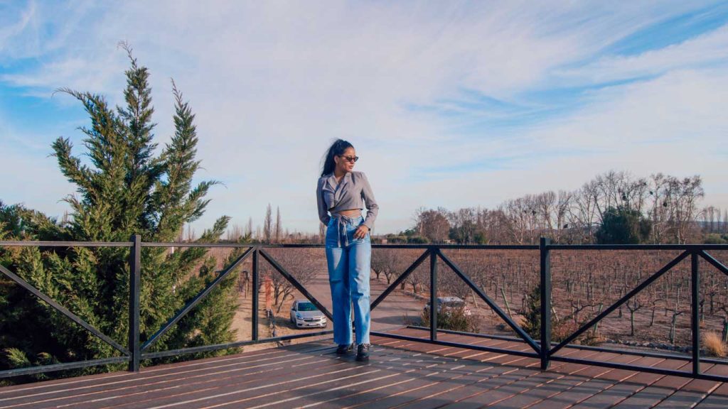 Mulher de óculos escuros, camisa cinza cropped e calça jeans posa em um deque de madeira com vista para vinhedos da vinícola Vistandes e árvores sem folhas, típicos do inverno, uma boa época para ir para Mendoza.