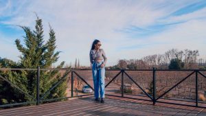 Mulher de óculos escuros, camisa cinza cropped e calça jeans posa em um deque de madeira com vista para vinhedos da vinícola Vistandes e árvores sem folhas, típicos do inverno, uma boa época para ir para Mendoza.