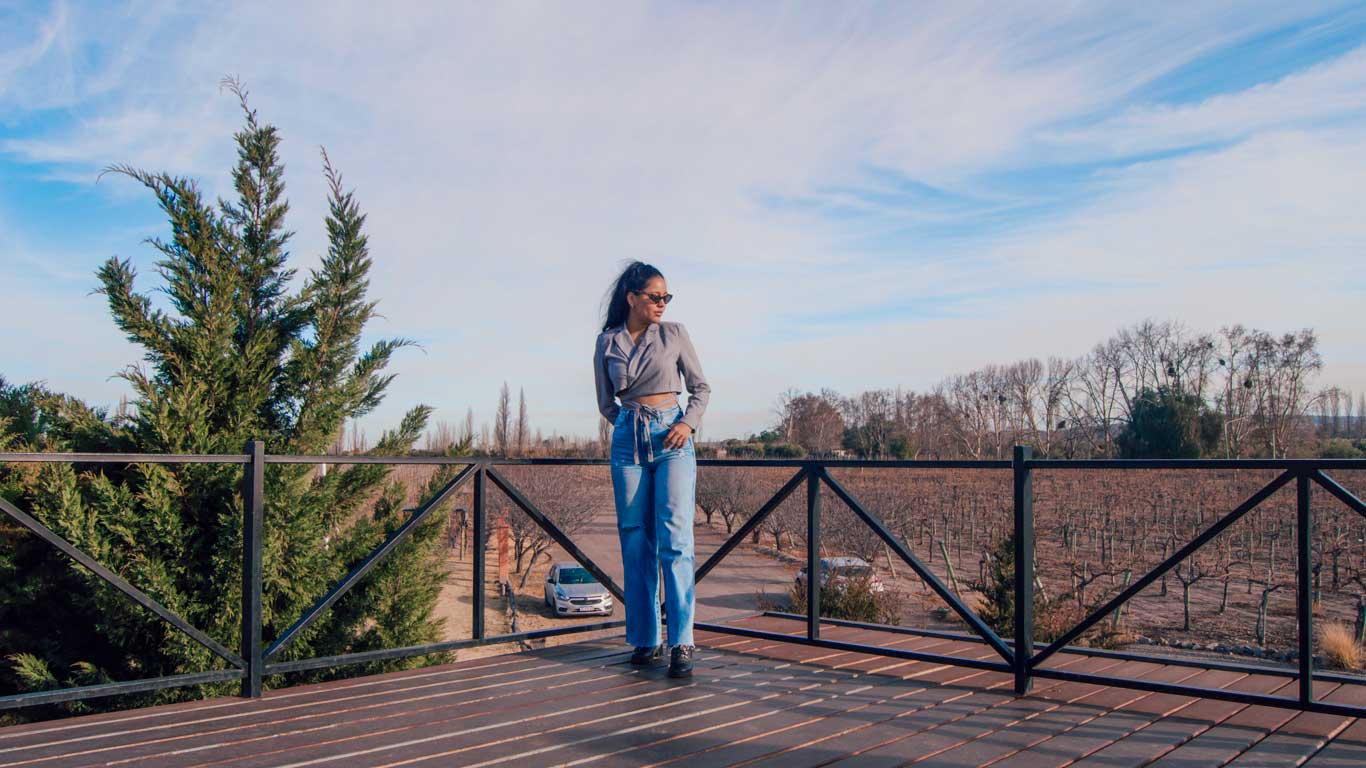 Mulher de óculos escuros, camisa cinza cropped e calça jeans posa em um deque de madeira com vista para vinhedos da vinícola Vistandes e árvores sem folhas, típicos do inverno, uma boa época para ir para Mendoza.